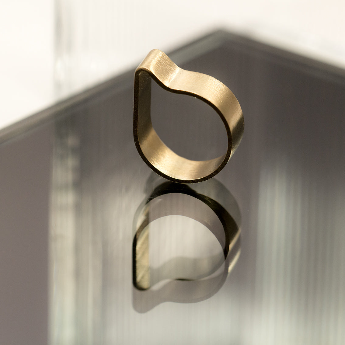 A minimally designed ring is sitting upright on a grey tinted mirror surface with a ribbed clear glass background piece. The reflection of the ring and the fluted glass are visible on the mirror base. The ring has a very simple design which seems like a brass band curved in the shape of a comma.