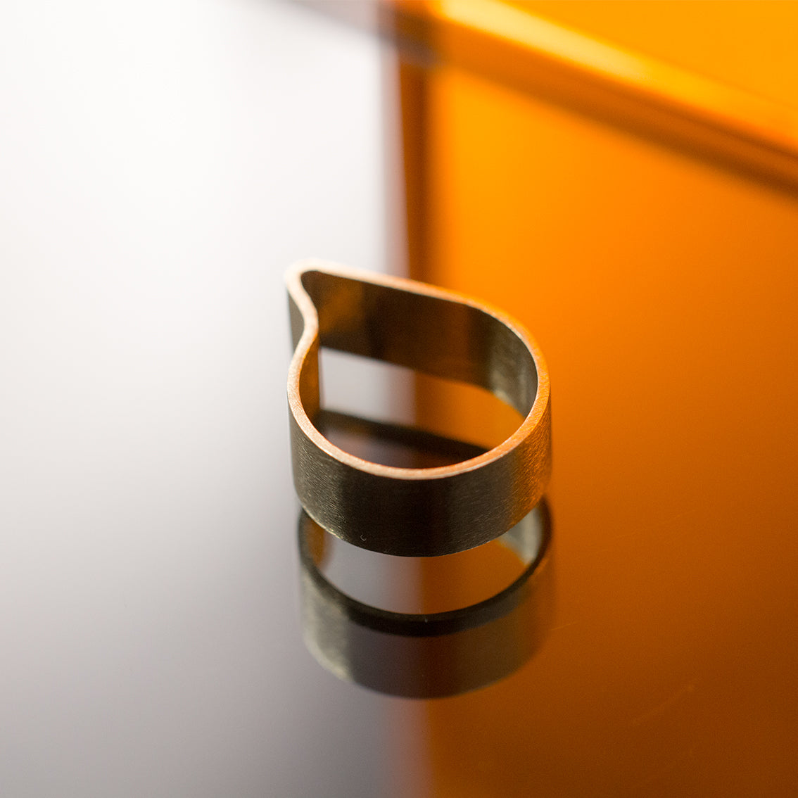 A minimally designed comma shaped ring with a soft curvy boundry. It's made of brushed brass and located on a reflective surface with orange coloured glass in the background. The ring and the orange tinted glass is reflected on the base reflective surface.