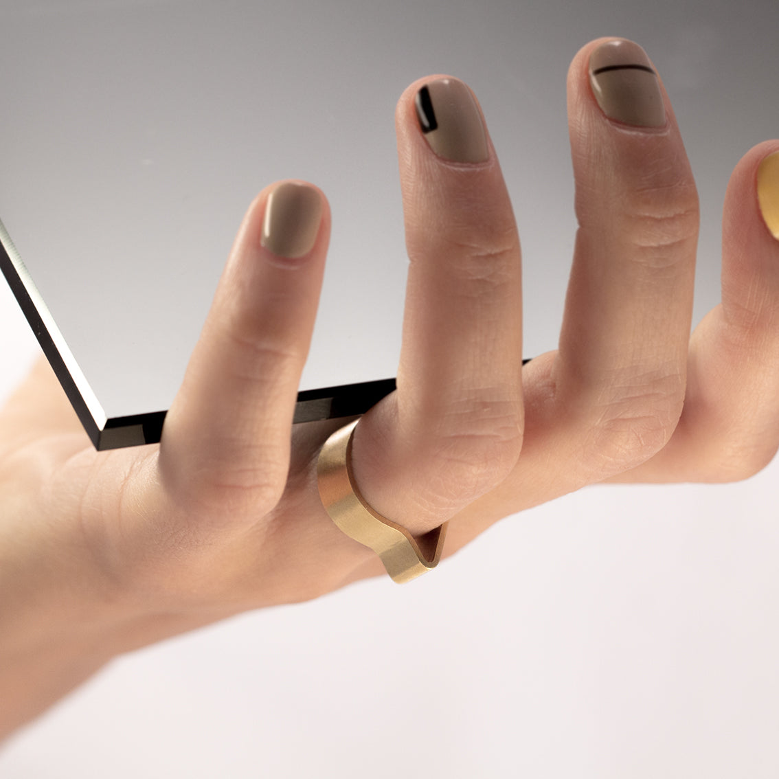 A right hand of a female person with trendy, interesting, graphical nail polish in light colours, holding a grey tinted mirror sample and wearing a feature ring. The ring has a very simple design which seems like a brass band curved in the shape of a comma. It's minimal in style and has architectural thinking in its' design.