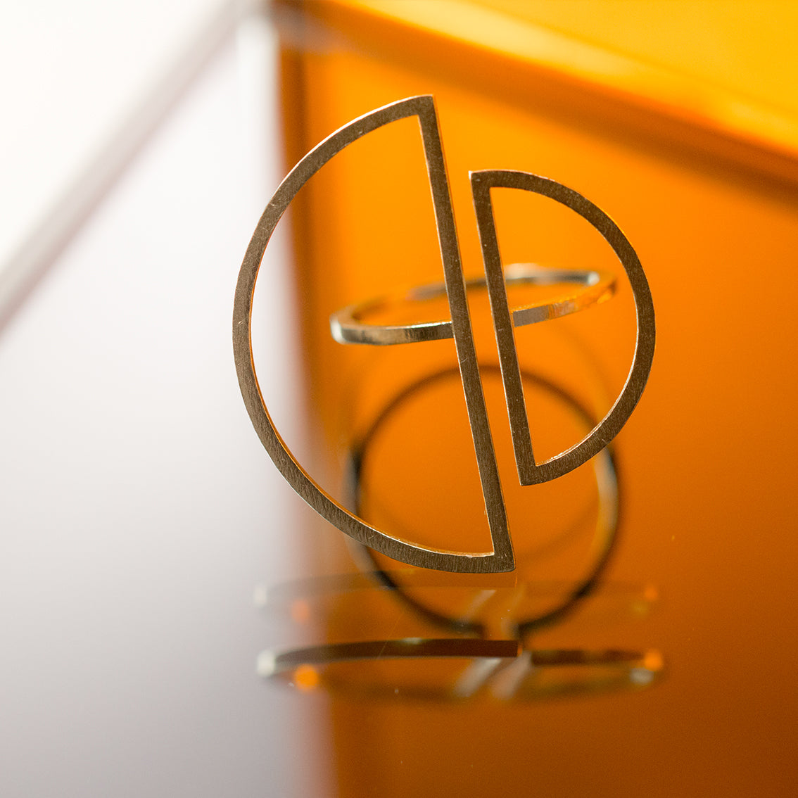 A minimally designed very light looking ring made of brass is sitting on a reflective surface with orange coloured glass in the background. The ring has a very thin band and is open on top to connect with two half circles which are also made of a very thin cross section. The two half circles are meant to hover on the adjacent fingers when the ring is worn.