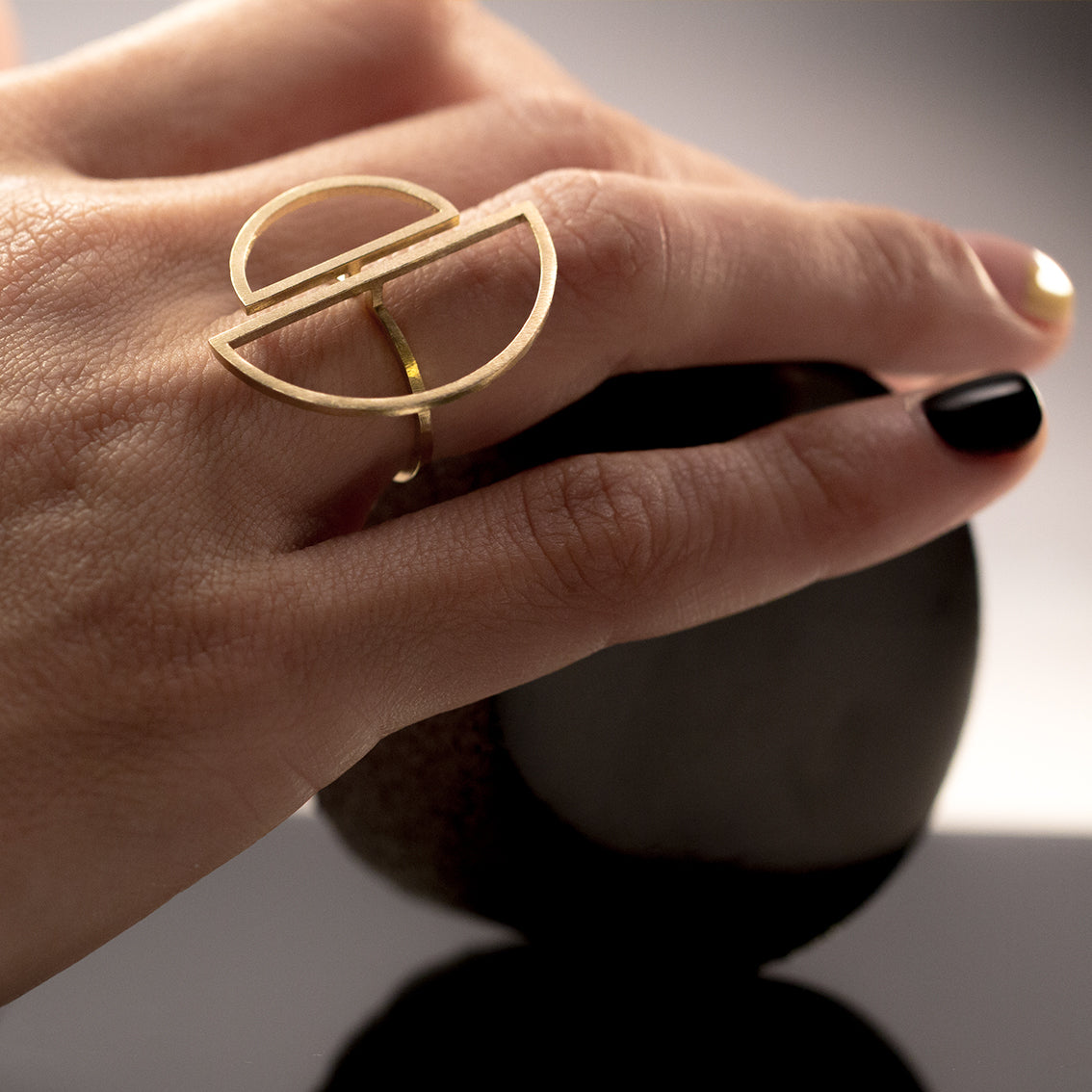 A right hand of a female person with trendy, interesting,  nail polish in black and light yellow, is lightly laying on a round black object ready to be rolled. The person is wearing a feature brass ring which glows lightly revealing it's minimal silhouette. The ring has a very thin band and is open on top to connect with two half circles which are also made of a very thin cross section. The two half circles are  hovering on the adjacent fingers.