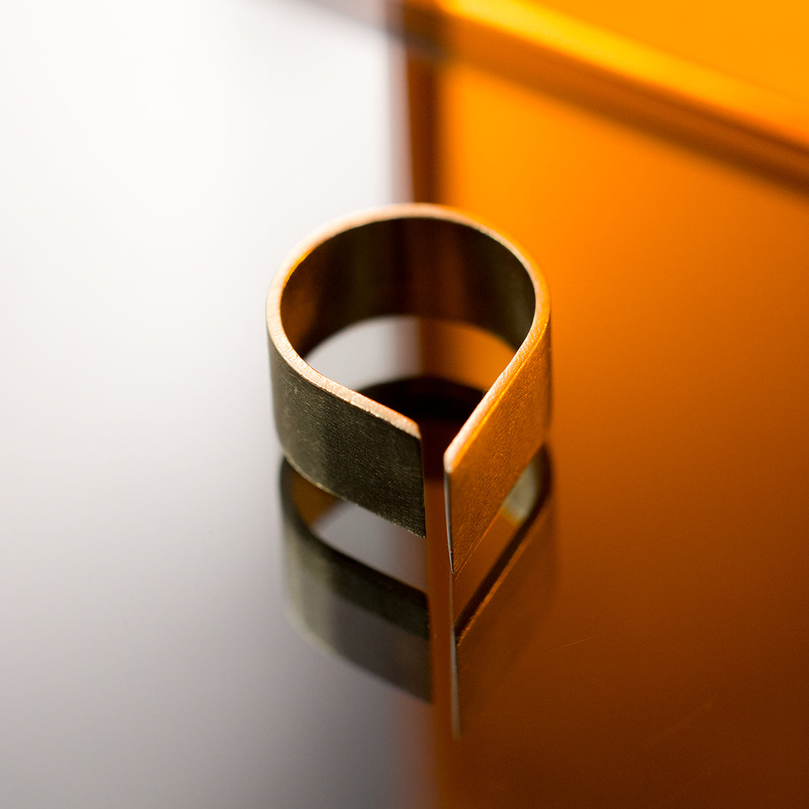 A shiny gold coloured ring with a very simple design is placed on a reflective surface. The ring is made of brushed brass and the shape is similar to a rain drop with a thin opening on top. The sharp edges of the 10mm thick band are catching the light softly to reveal the shape of the ring. There is an orange tinted glass behind and the reflection of this is also visible on the mirror surface.