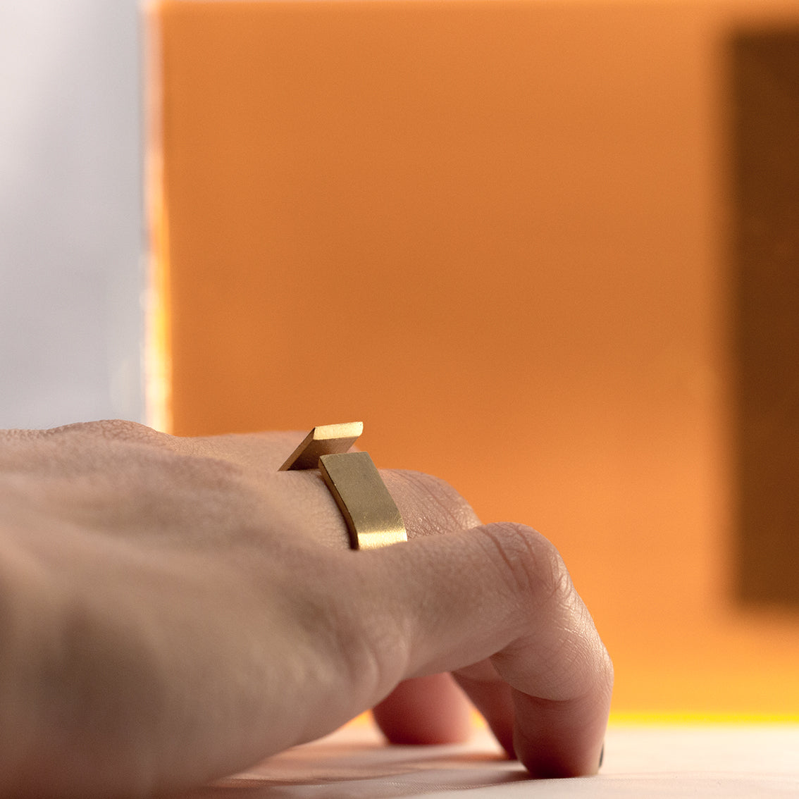 A right hand of a female person with a feature ring on its ring finger is lightly laying on an off white surface. The photo is taken from behind leveled with the height of the hand. The statement ring with sharp edges is the focus of the photo. The ring is made of brushed brass and the shape is similar to a rain drop with a thin opening on top. The sharp edges of the 10mm thick band are catching the light softly to reveal the shape of the ring. 