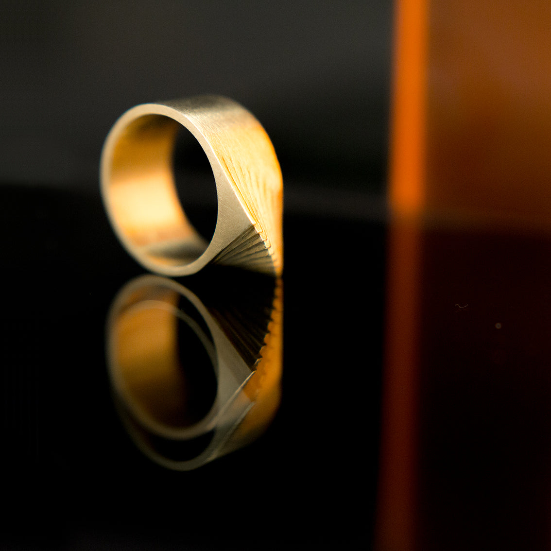 A photo of an attention grabbing brass ring laying on a reflective black surface with some orange coloured reflection in the background. The photo has dimmed lighting and the ring has a low glow. The ring seems to be made of layers rotating on the same axes. It has a pointy top edge which seems like a spine of a creature. The rhythm of the layers of the ring is the focus of the photo. The photo has sexy elements because of shapes, reflections, colour and dimmed lighting. 