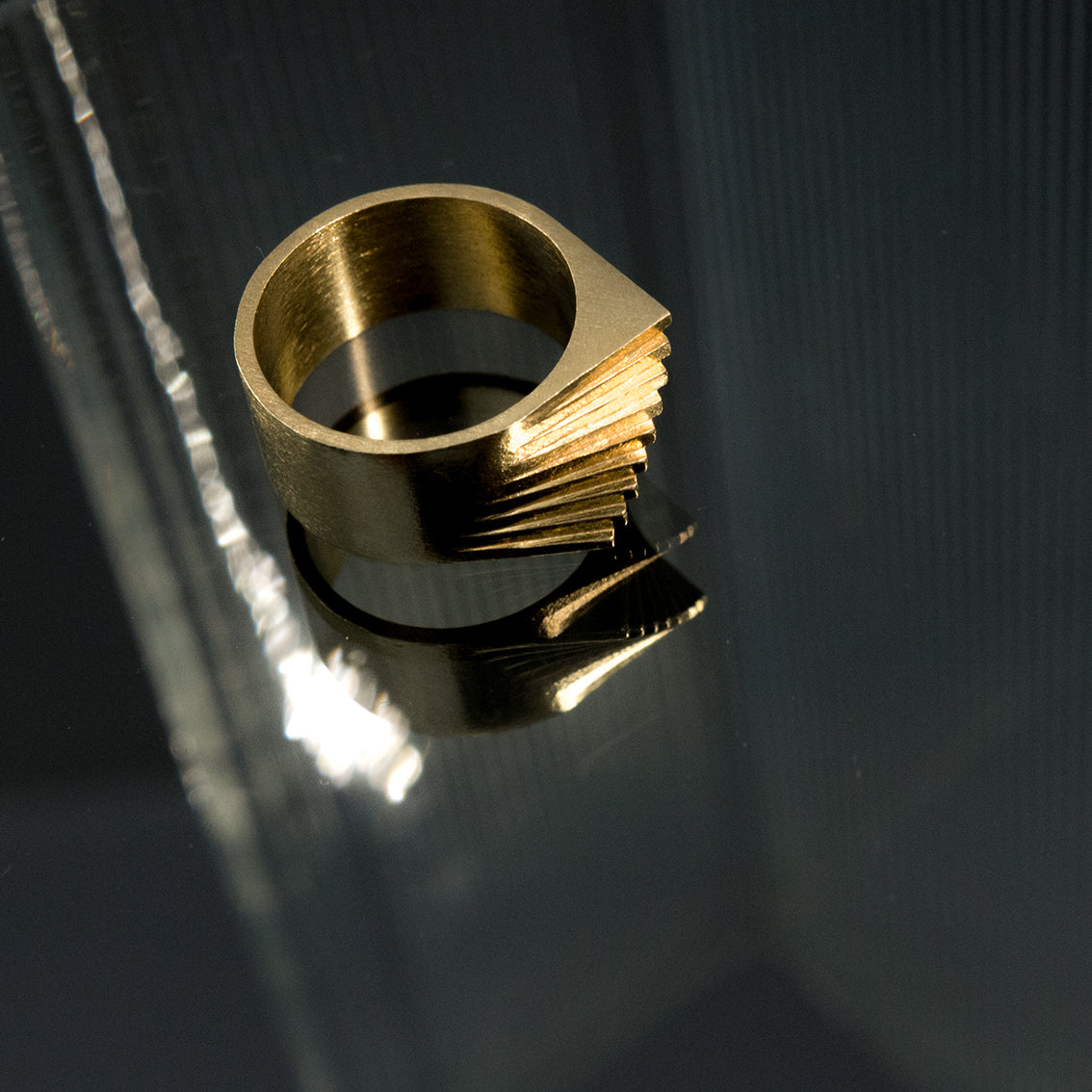 A photo of an attention grabbing brushed brass ring laying on a black tinted mirror. The reflection of the ring and ribbed glass is visible on the mirror. The photo has dimmed lighting and the ring has a low glow. The ring seems to be made of layers rotating on the same axes. It has a pointy top edge which seems like spiral stairs. The rhythm of the layers of the ring is the focus of the photo. The photo has sexy elements because of shapes, reflections, colour and dimmed lighting.