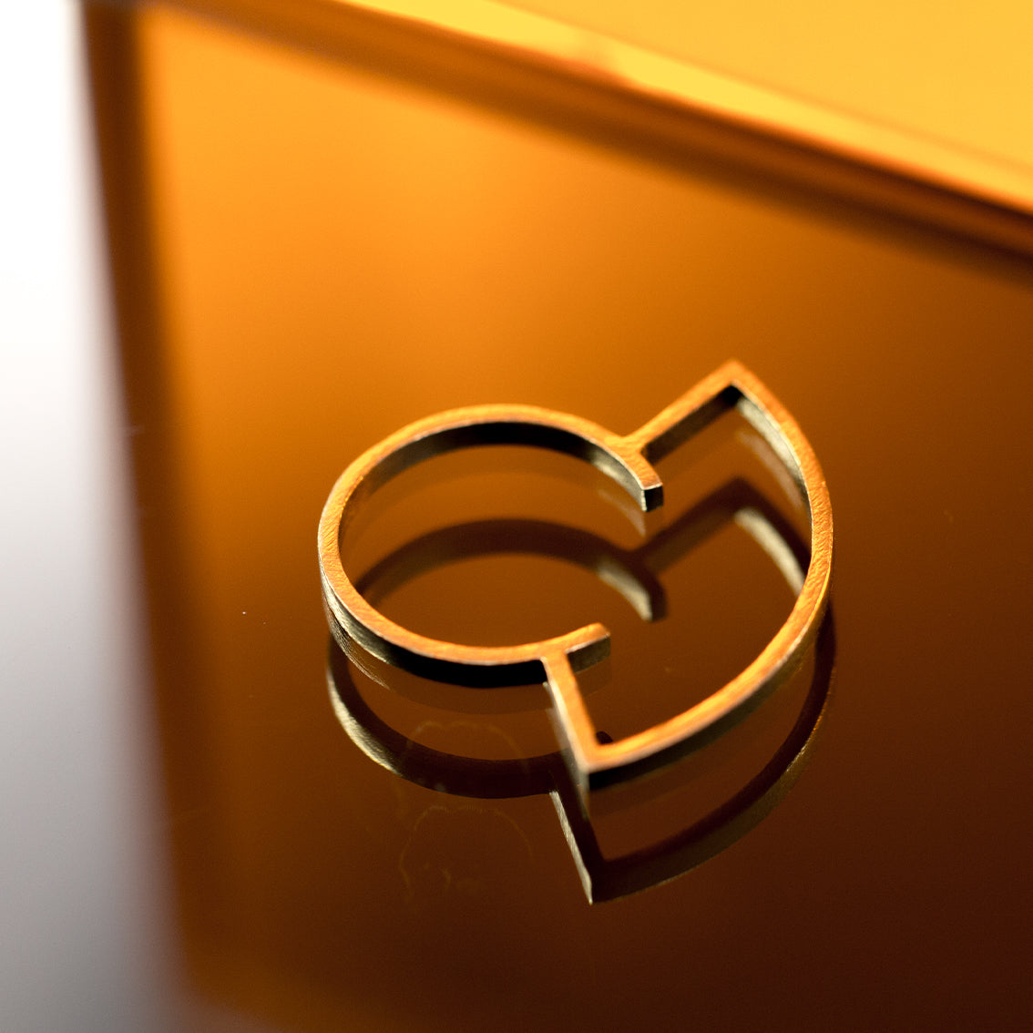 A gold coloured brass ring on an orange background which is designed as a silhouette and has a very thin band. It's very light, contemporary, playful yet an elegant design.