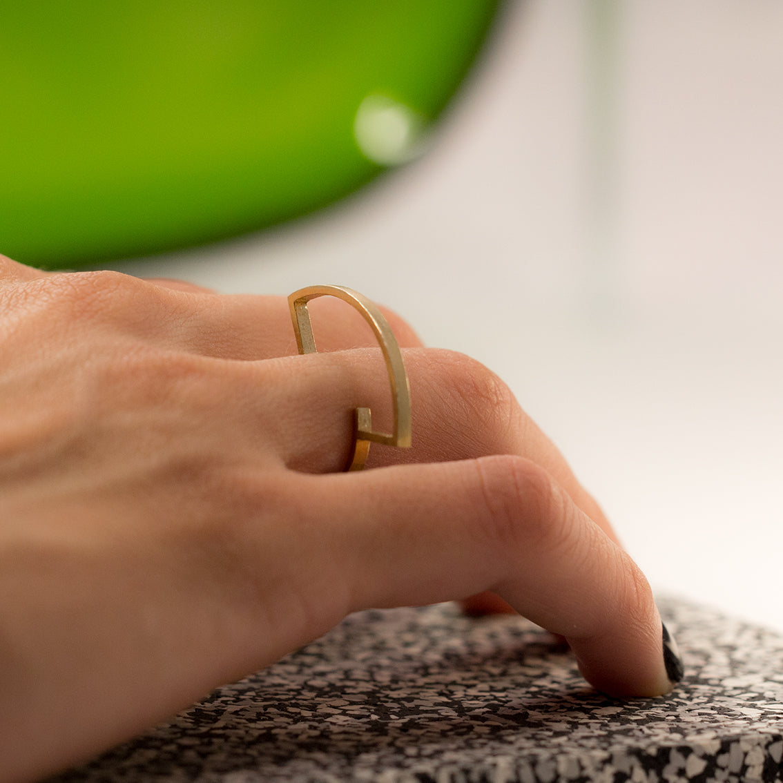 A photo of a right hand from the back with a minimal, geometric brass ring on the ring finger. The hand is lightly laying on a white and grey speckled black rubber surface – a rubber flooring sample for gyms. There is also a green shiny object in the background which makes the photo obtain a very joyful vibe.  The statement ring with sharp edges is made of brushed brass and has a very thin band. It's designed as a silhouette that reminds a totem. It's very light, contemporary, playful yet an elegant design.