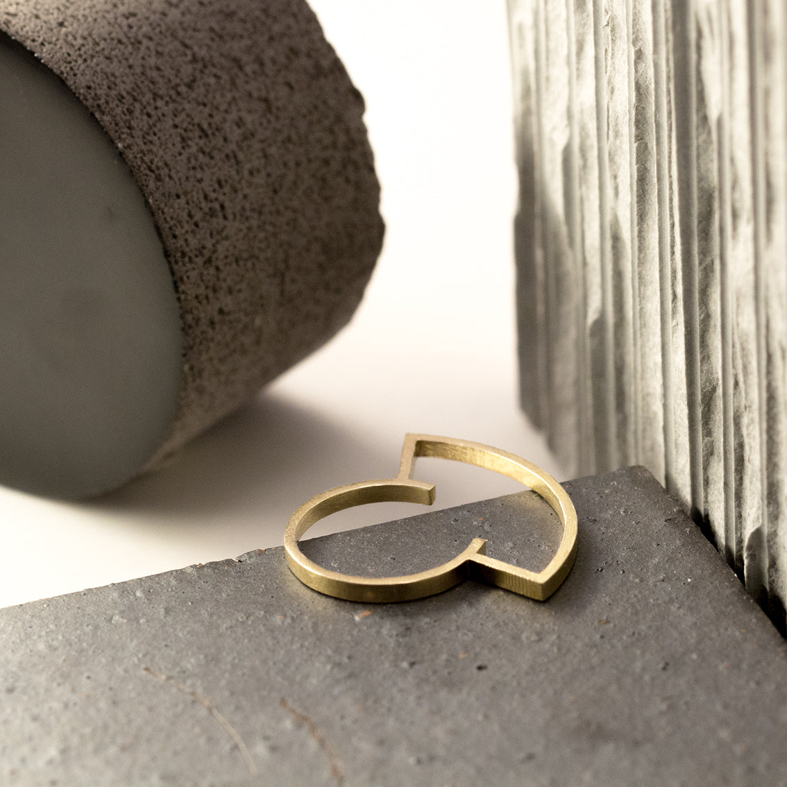 A brass ring which is designed as a silhouette and has a very thin band. It's very light, contemporary, playful yet an elegant design. It’s sitting among architectural materials including grey stone, concrete, lava stone. 